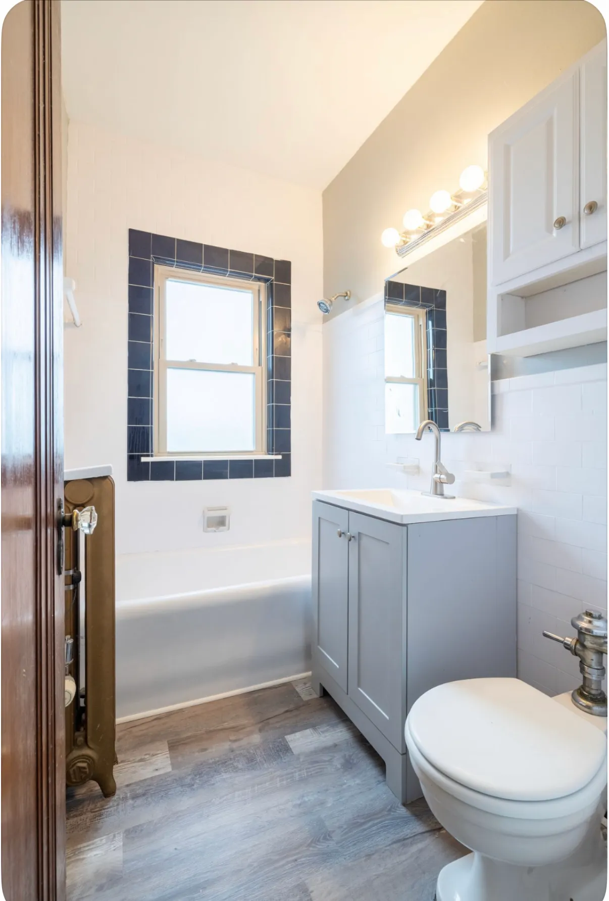 The same bathroom after work: navy tiled window surround, grey vanity and new flooring, with the original cast iron radiator and wood door kept